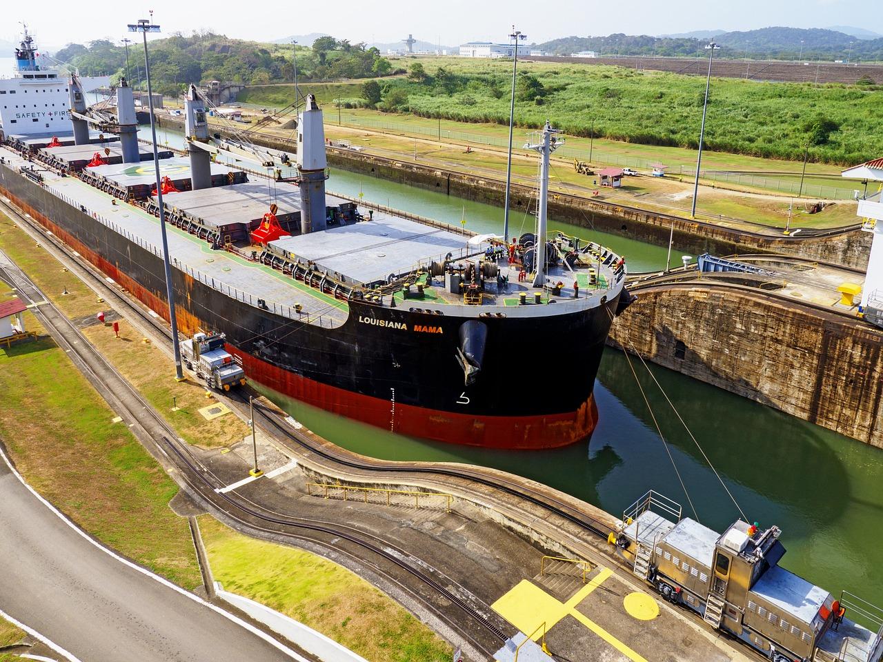 A large cargo ship navigates through the Panama Canal, flanked by towering concrete walls and lush greenery. Tugboats guide the vessel as it moves steadily through the narrow lock, with ropes securing its path. In the background, mechanical gates and lock machinery operate to regulate the water level, enabling the ship’s smooth transit across this engineering marvel connecting the Atlantic and Pacific Oceans.